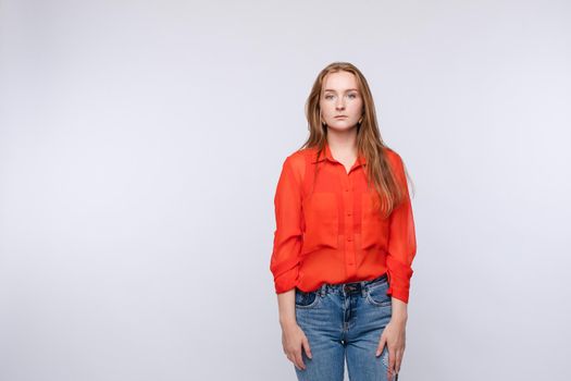 Cropped view of upset young woman wearing red blouse and jeans standing on isolated background. Calm female adult looking at camera and posing in studio. Concept of sadness and copy space.