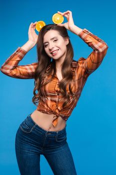 Young girl in casual clothes hiding her face behind fruits. Brunette lady in checkered shirt and jeans holding halves of oranges as sunglasses. Beatiful cute woman with opened belly happily smiling.