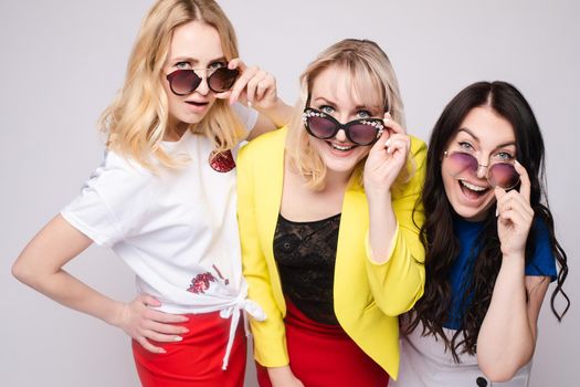 Stock portrait of three women in fashionable sunglasses looking at camera with different facial expressions. Women looking at camera over their sunglasses with excitement and suspense.