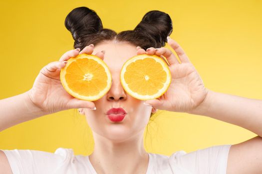 Studio headshot of young funny brunette with hairstyle and red lips holding halved oranges on eyes against bright yellow background.