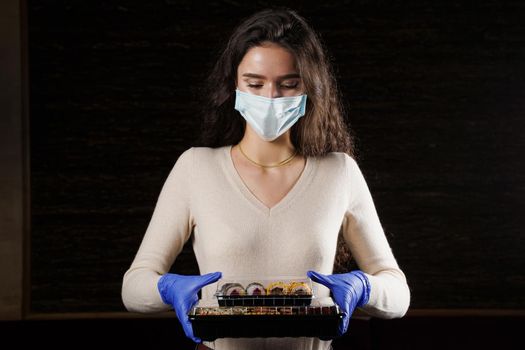 Girl with sushi. Food courier in medical mask. Delivery at quarantine coronavirus covid-19. Young woman holding 2 sushi boxes in hands using gloves