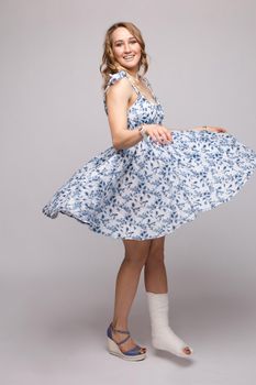 Full length portrait of positive beautiful lady in summer dress and sandals dancing with her skirt flying with plaster on her broken leg. Isolate on grey. Smiling woman dancing with plaster.