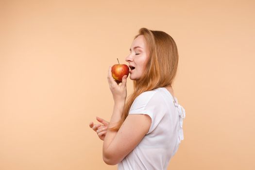 Beautiful young girl with tied on back hair eating tasty apple on lunch. Side view of attractive model promoting healthy lifestyle. Brunette woman with white teeth holding delicious fruit in her hand.