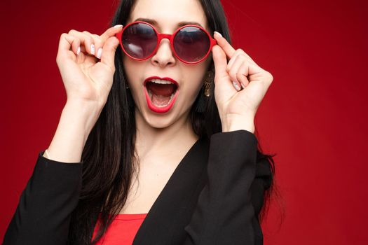 Studio headshot of funny cheerful and positive brunette woman with red lips showing or expressing happiness and excitement in fashionable red round sunglasses. She is posing against red background.