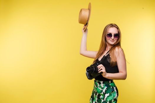 Front view of bright young girl wearing straw hat and glasses posing on yellow isolated background in studio. Female tourist keeping camera, looking at camera and smiling. Concept of summer.