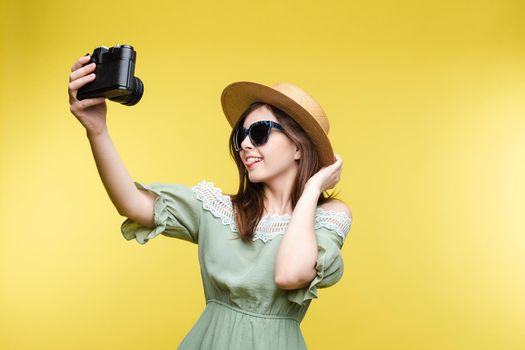 Studio portrait of fashionable young brunette girl in summer dress and stylish hat and sunglasses posing while taking self-portrait via film camera. Isolate on yellow.