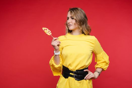 Studio portrait of confused brunette in fashionable summer yellow dress with belt looking at sweet lollipop with suspicion. Isolate on red background. She wants sweet but cannot eat it.