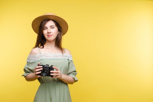 Front view of bright young girl wearing straw hat and glasses posing on yellow isolated background in studio. Female tourist keeping camera, looking at camera and smiling. Concept of summer.