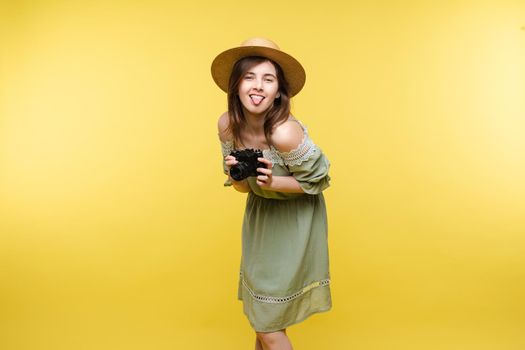 Front view of bright young girl wearing straw hat and glasses posing on yellow isolated background in studio. Female tourist keeping camera, looking at camera and smiling. Concept of summer.