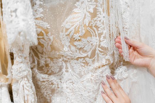 Bride touches luxurious white cream wedding dress on hangers. Dresses made of silk chiffon, tulle and lace. Luxury pearls and crystals pendants on the sleeves of a delicate color of a wedding dress