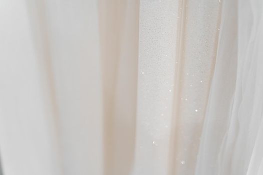 Cloth of wedding dresses made of silk chiffon, tulle and lace. Beautiful White cream bridal dress on hangers in wedding salon. Pearls and crystals pendants on the sleeves of a wedding dress