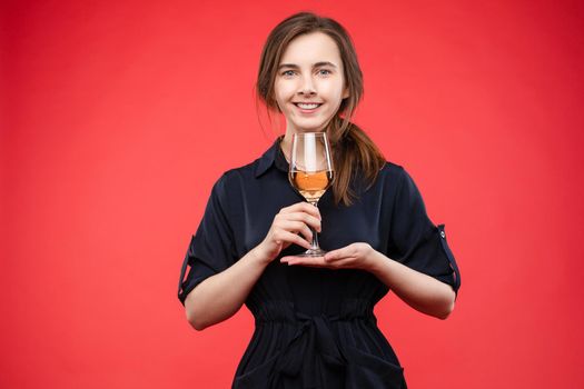 Young pretty lady in casual clothes holding delicious drink in hands. Brunette happy girl enjoying her sweet beverage and smiling. Beautiful woman in black dress standing with glass of expensive wine.