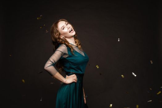 Happy young girl looking surprisingly while golden confetti falling. Beautiful woman in emerald elegant long dress smiling and dancing among decorations. Gorgeous lady celebrating birthday on party.