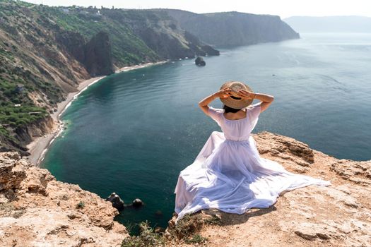 Portrait of a young woman on the beach by the sea, sitting with raised hands in a white dress and hat. Back photo.