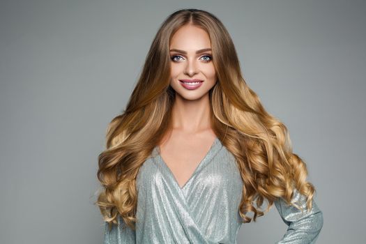 Studio portrait of attractive blonde woman with long thick hair and professional make-up wearing elegant silver blouse smiling at camera over grey background. Isolate. Beauty concept.