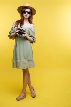 Front view of bright young girl wearing straw hat and glasses posing on yellow isolated background in studio. Female tourist keeping camera, looking at camera and smiling. Concept of summer.