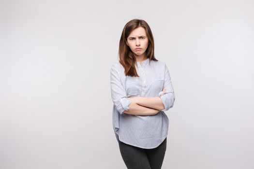 Studio portrait of disappointed young girl in striped casual shirt and black trousers looking at camera and frowning her eyebrows with folded arms on chest.