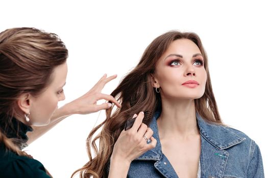 View from side of professional stylist doing hairstyle to young model in studio. Attractive woman in denim jacket standing and posing on white isolated background. Concept of beauty and fashion.