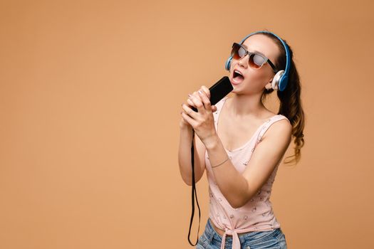 Happy young woman wearing headphones listening music and dancing on isolated background. Cheerful girl in glasses keeping microphone and singing in studio. Concept of relax and fun.