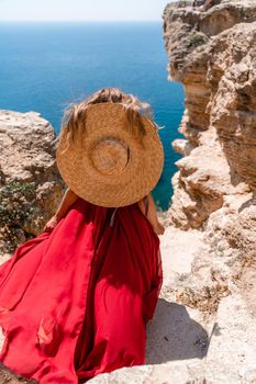 A girl with loose hair in a long red dress descends the stairs between the yellow rocks overlooking the sea. A rock can be seen in the sea. Sunny path on the sea from the rising sun.