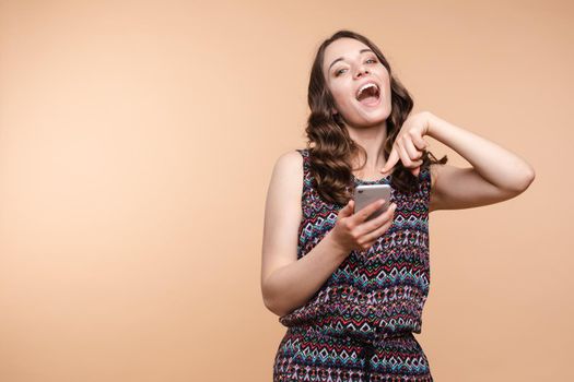 Studio portrait of beautiful caucasian brunette woman in patterned overall pointing at her smartphone with index finger. She is certain or sure about something. I know gesture.