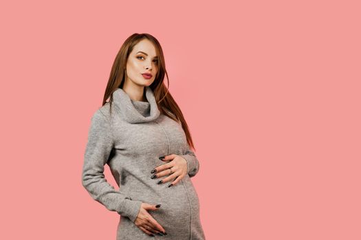 Pregnancy of a happy beautiful girl in a gray sweater on a pink background. Pregnant woman. Expectation of the child. Maternity leave