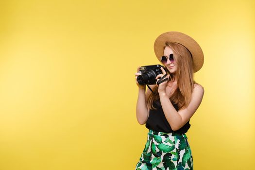 Studio portrait of pretty smiling young woman with long red hair in summer clothes and a hat with sunglasses taking a photo with professional photo camera. She is isolate on yellow background.