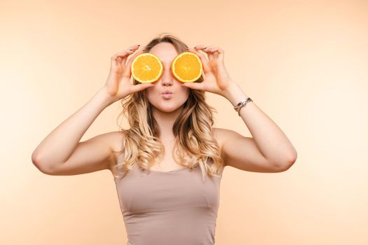 Front view of cheerful young blonde posing with fresh oranges on isolated background. Funny girl keeping fruit and closing eyes in studio. Concept of happiness and health.