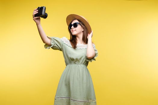 Happy traveler woman posing making selfie using camera isolated at yellow studio background medium long shot. Smiling fashionable young girl wearing straw hat and sunglasses having positive emotion