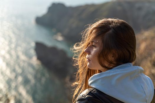 Woman tourist enjoying the sunset over the sea mountain landscape. Sits outdoors on a rock above the sea. She is wearing jeans, a blue hoodie and a black leather jacket. Healthy lifestyle, harmony and meditation.