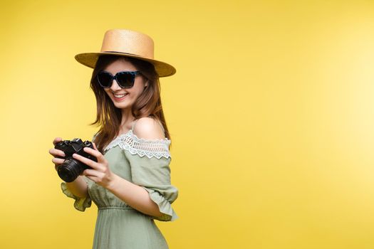 Front view of bright young girl wearing straw hat and glasses posing on yellow isolated background in studio. Female tourist keeping camera, looking at camera and smiling. Concept of summer.