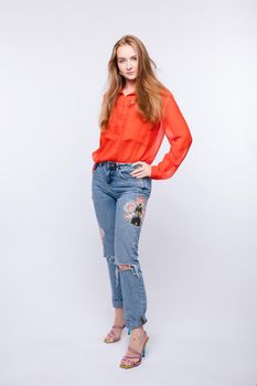 Side view of young pretty girl wearing red blouse and jeans posing on grey isolated background. Longhaired female model standing and looking at camera in studio. Concept of full length.