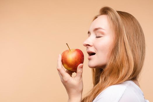 Beautiful young girl with tied on back hair eating tasty apple on lunch. Side view of attractive model promoting healthy lifestyle. Brunette woman with white teeth holding delicious fruit in her hand.