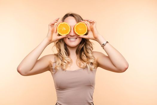 Front view of cheerful young blonde posing with fresh oranges on isolated background. Funny girl keeping fruit and closing eyes in studio. Concept of happiness and health.