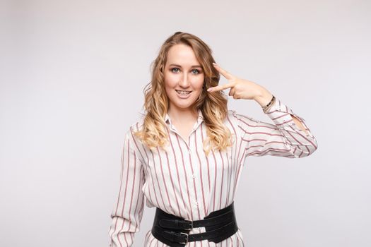 Studio portrait of pretty young Caucasian woman in striped shirt with fashionable wide waist belt showing peace sign near her face and smiling at camera. Isolate on white.