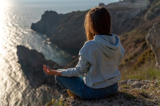 Woman tourist enjoying the sunset over the sea mountain landscape. Sits outdoors on a rock above the sea. She is wearing jeans and a blue hoodie. Healthy lifestyle, harmony and meditation.