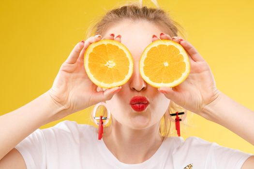 Studio headshot of young funny brunette with hairstyle and red lips holding halved oranges on eyes against bright yellow background.