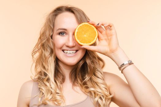 Studio headshot of young funny brunette with hairstyle and red lips holding halved oranges on eyes against bright yellow background.