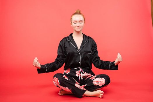 Young girl in black pijama sitting on floor and meditating with closed eyes. Beautiful happy woman in casual clothes doing yoga and practicing breathing. Pretty relaxed lady caring about her health.