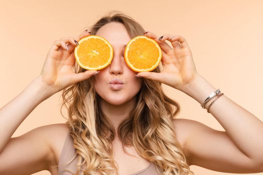 Front view of cheerful young blonde posing with fresh oranges on isolated background. Funny girl keeping fruit and closing eyes in studio. Concept of happiness and health.