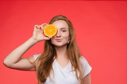 Studio headshot of young funny brunette with hairstyle and red lips holding halved oranges on eyes against bright yellow background.