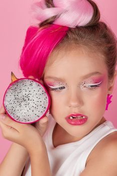 Close-up portrait of pretty girl with pink hairstyle with dragon fruit on pink background. Studio shot of charming tween girl with pink make up enjoying juicy red pitaya. exotic Pitahaya fruit.