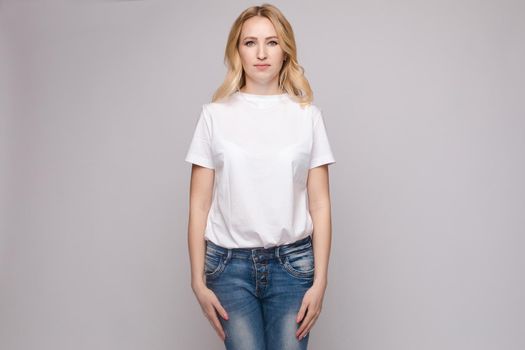 View from front of beautiful slim woman wearing white shirt and jeans standing steady on frey isolated background. Young blonde looking at camera, smiling and posing in studio.