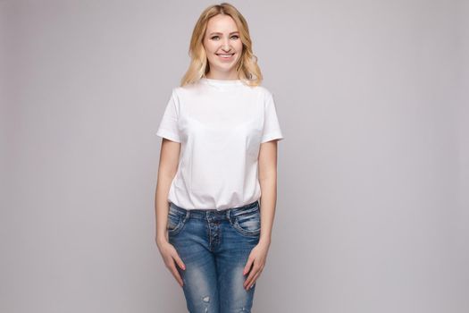 View from front of beautiful slim woman wearing white shirt and jeans standing steady on frey isolated background. Young blonde looking at camera, smiling and posing in studio.