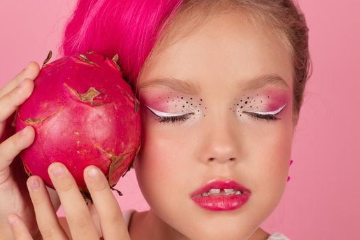 Close-up portrait of pretty girl with pink hairstyle with dragon fruit on pink background. Studio shot of charming tween girl with pink make up enjoying juicy red pitaya. exotic Pitahaya fruit.