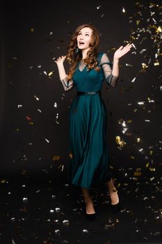 Happy young girl looking surprisingly while golden confetti falling. Beautiful woman in emerald elegant long dress smiling and dancing among decorations. Gorgeous lady celebrating birthday on party.