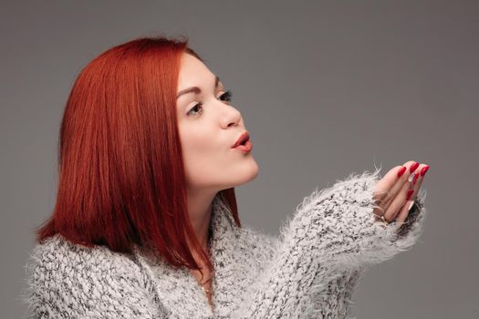 Beautiful young girl with red hair and red nails holding her hands together and blowing on white lightening ball. Pretty woman in grey sweater catching star and making wishes when snowing.