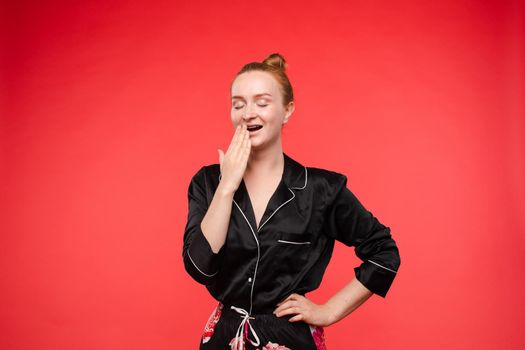 Front view of tired woman wearing black nightwear yawning on red isolated background. Redhaired girl preparing for night time and bedding. Concept of sleeping, dreaming and relaxing.