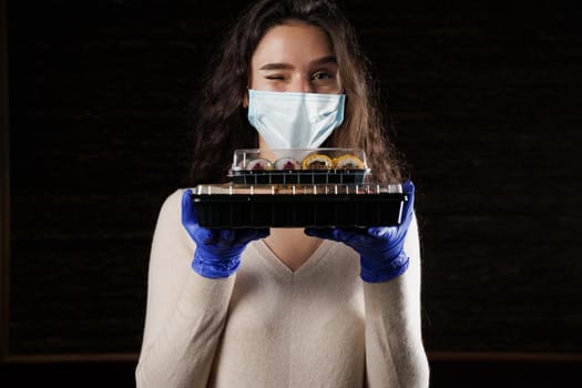 Girl with sushi. Food courier in medical mask. Delivery at quarantine coronavirus covid-19. Young woman holding 2 sushi boxes in hands using gloves