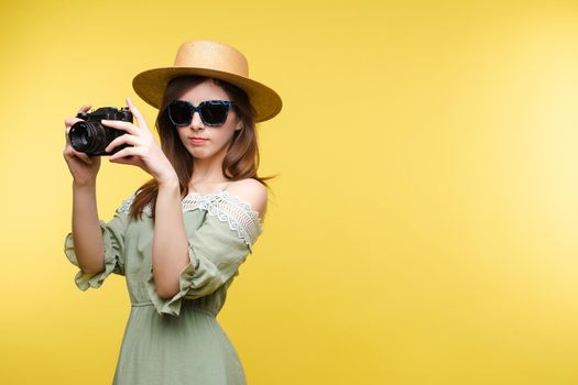 Front view of bright young girl wearing straw hat and glasses posing on yellow isolated background in studio. Female tourist keeping camera, looking at camera and smiling. Concept of summer.